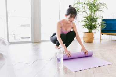 Genç Asyalı kadın, yoga yapmaya hazırlanırken yoga minderini su şişesiyle yuvarlıyor. Evde ya da yoga stüdyosunda çalışıyorum. Sağlıklı alışkanlıklar, formda kalma, kilo verme kavramları.