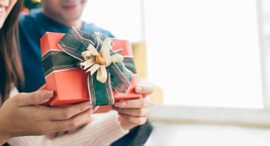 Yakın plan genç Asyalı bir kadın elinde kırmızı bir kutuda erkek arkadaşıyla birlikte arka planda süslenmiş bir Noel ağacı olan bir kurdeleyle Noel hediyesi tutuyor..