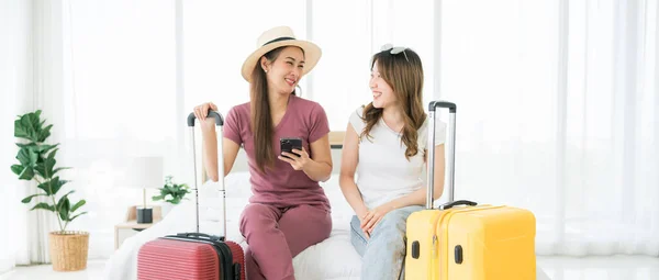 两名快乐的亚洲女人用智能手机预订了一间酒店房间 并用信用卡付帐 她们一起坐在床上带着行李 计划度假旅行 带有复制空间的图像 — 图库照片