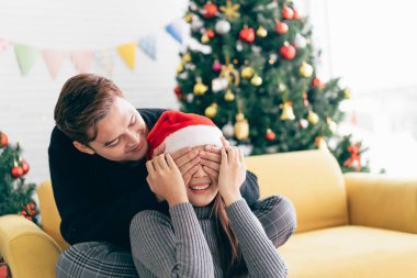 Genç Asyalı adam sürpriz yapar ve arka planda bir Noel ağacı ile evde bir Noel hediyesiyle gözlerini kapatır. Kopyalama alanı olan resim.