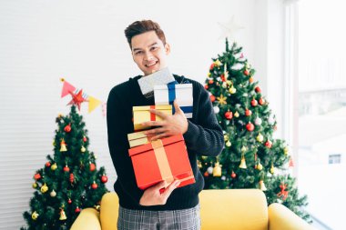 Heyecanlı ve şaşırmış Asyalı genç bir adamın portresi. Elinde, arka planda süslenmiş Noel ağacıyla oturma odasında duran hediyeler var..