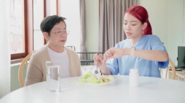 Asian woman caregiver wearing medical scrub takes care of senior Asian woman by feeding vegetarian salad to eat at home. caregivers visit home Home health care concept and nursing home.