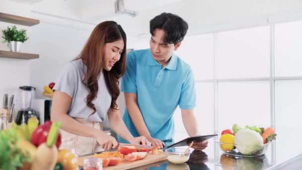 幸せなアジアのカップル健康的な料理を調理します 女性の間にレシピを検索し チェックするためにデジタルタブレットを使用して男は自宅のキッチンで新鮮な野菜とベジタリアンの健康的なサラダを準備します — ストック動画