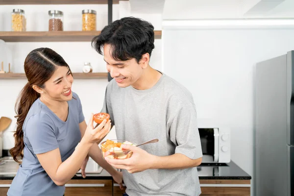 Healthy young Asian couple feeding a fresh sliced tomato together with a smile and happy emotion. An active lifestyle with healthy and vegetarian food advertisement. Image with copy space.