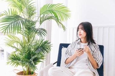 Genç, güzel bir kadın gülümser ve yeşil bir bitkiyle modern yaşam alanının sandalyesine oturur, bir fincan sıcak kahve içerek rahatlar ve rahat bir evde sakin vakit geçirir ve ev ortamının tadını çıkarır..