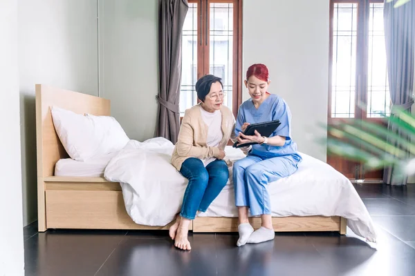 Asian woman therapy nurse wearing scrubs report the health status of senior Asian woman with digital tablet in the bedroom. Caregiver visit at home. Home health care and nursing home concept.