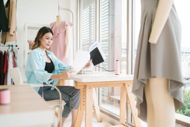 Kendine güvenen Asyalı bir terzinin portresi terzi atölyesinde giysiler üzerinde çalışıyor. Çekici bir bayan moda tasarımcısı gülümseyip arka planda elbise mankeni olan kameraya bakar..