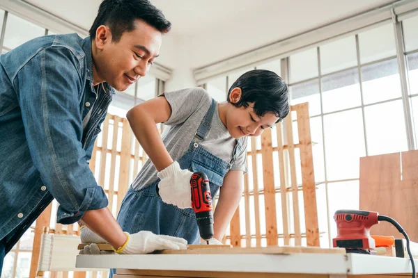 Asian father and son work as a woodworker or carpenter, Father teaches his son to drill holes in a wooden plank carefully together with teamwork. Craftsman carpentry working at home workshop studio.