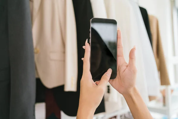 A close up woman hands chooses clothes on the rail by taking a photo with smart phone for making a decision to buy or sharing on social media in a clothing store. Clothes retail shopping concept.