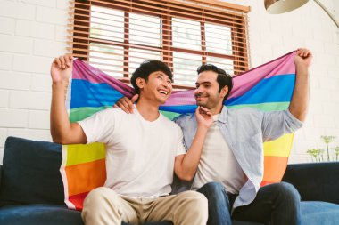 Happy caucasian and Asian LGBT couple, sitting on the sofa holding and waving rainbow LGBT Pride flag together in the living room at home. Diversity of LGBT relationships. A gay couple concept.