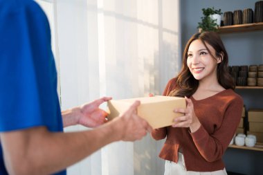 Asyalı bir kadın müşteri, üniformalı beyaz bir kuryeden, evdeki giriş kapısının önüne teslim etmesi için bir paket nakliye paketi almış. Ev paketi teslimat hizmeti kavramı.