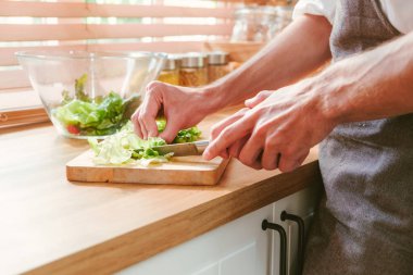 Close-up hand of caucasian gay couple cooking salad by using a knife to cut lettuce on the cutting board over the kitchen counter at home with morning light. LGBT relationships. Gay couple concept