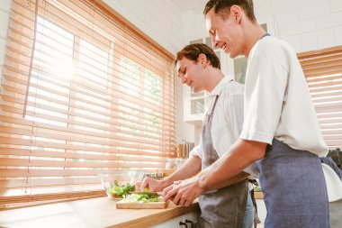 Happy caucasian gay couple making salad together One person used a knife to cut lettuce in front. And another person watching from behind in the kitchen at home. LGBT relationships. Gay couple concept