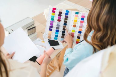 Two Asian female fashion designers or dressmakers brainstormed and discuss the design of the new collection with samples of fabrics and sketches on the tablet. Working together in the studio.