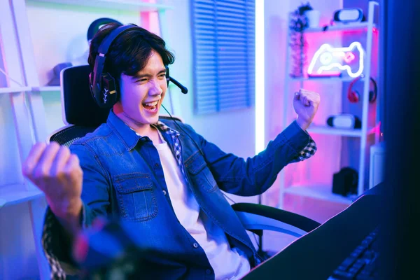 Asian male pro gamer winning an esports game with victory emotion while sitting on the gamer chair, wearing headphones with a microphone. The player rejoices in victory in the competition.