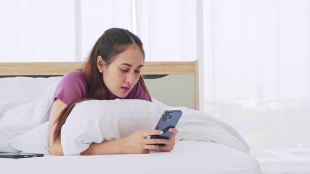 幸せなアジアの女性は 午前中に指で画面に触れるスマートフォンを使用してベッドに横たわっていた 女性はスマートフォンからのソーシャルネットワーク チャットやオンラインショッピングのためのデバイスを使用します — ストック動画