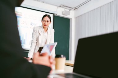 A cheerful and confident Asian businesswoman stands, present bar charts data from projector screen to her office colleagues. Asian business women leader role at the meeting.