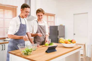 Önlük takan mutlu beyaz eşcinsel çiftin portresi, birbirlerine gülümseyerek bakıyorlar ve evdeki mutfakta birlikte yemek pişirmekten zevk alıyorlar. LGBT ilişkileri. Eşcinsel çift yemek pişirme konsepti.