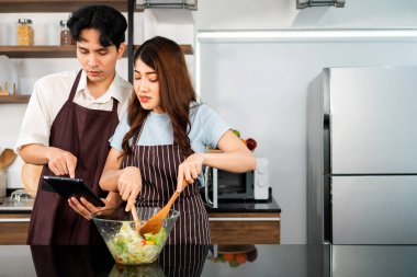 Önlük giyen mutlu Asyalı çift. Bir kadın, tableti işaret ederken bir erkek ev mutfağında havuç, domates ve yeşil meşe gibi taze sebzelerle sağlıklı bir vejetaryen salatası hazırlar..