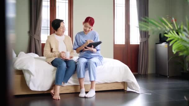 スクラブを装着したアジア系女性看護師は ベッドルームにタブレットを装着したアジア系シニア女性の健康状態を報告しています 介護者が家に来る ホームヘルスケアと介護ホームコンセプト — ストック動画