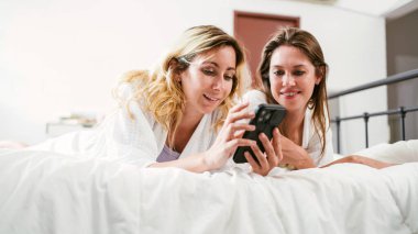 Beyaz bornozlu, mutlu iki arkadaş. Evde yatakta yatarken birlikte akıllı telefon kullanıyor ve gülümsüyor. LGBTQ çiftler için spa ve tedavi aktiviteleri.