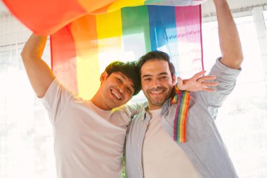 Mutlu Kafkasyalı ve Asyalı LGBT çifti, LGBT gurur bayrağının altında ayakta ve el sallıyorlar evdeki oturma odasında. LGBT ilişkilerinin çeşitliliği. Eşcinsel çift konsepti.