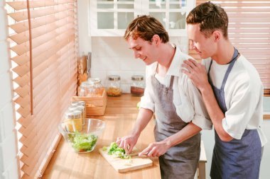 Happy caucasian gay couple making salad together One person used a knife to cut lettuce in front. And another person watching from behind in the kitchen at home. LGBT relationships. Gay couple concept