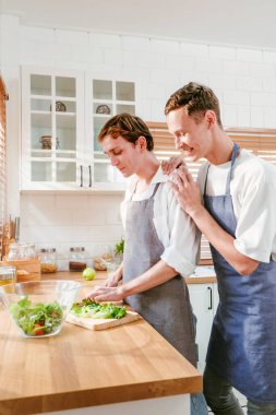 Happy caucasian gay couple making salad together One person used a knife to cut lettuce in front. And another person watching from behind in the kitchen at home. LGBT relationships. Gay couple concept