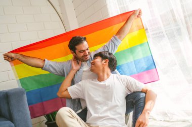 Happy caucasian and Asian LGBT couple, sitting on the sofa holding and waving rainbow LGBT Pride flag together in the living room at home. Diversity of LGBT relationships. A gay couple concept.