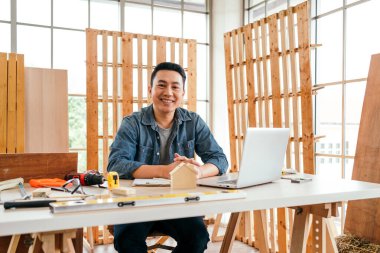 Portrait of a smile Asian male father sitting and working with a laptop on the table with the instrument of measure and working tools on the table at home woodwork studio.