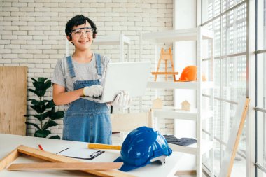 A cheerful Asian carpentry boy wearing safety goggles and work gloves, working on a laptop in the home workshop studio. Construction work and repairs. Carpentry and family concept.