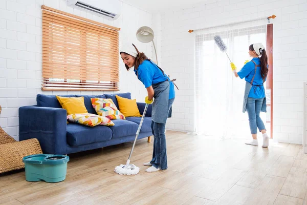 两名亚洲年轻的专业清洁服务女工团队在家里工作 女管家用羽毛抹布擦地板上的湿湿的拖把 另一个用羽毛抹布擦窗帘 图库照片