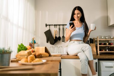 Kafkasyalı çekici bir kadın, rahatlarken, kahve içerken ve sabahları evdeki mutfak tezgahında otururken akıllı telefon kullanıyor. Mutfakta cep telefonuyla alışveriş yapan mutlu bir kadın..