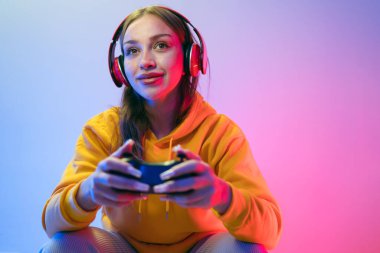 A young Caucasian beautiful woman in a yellow hoodie playing a video game using a joystick and headphones with fun, focus, surprise, and a winning victory face with blue and red neon light. clipart