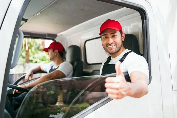 Happy professional truck driver with his assistant wearing a red cap thumb up, smiling, looking at the camera from a truck window before delivering parcel. A truck driver and delivery service concept.