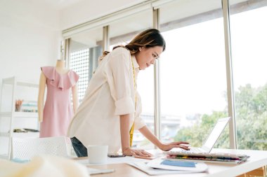 Kendine güvenen Asyalı terzi kadın terzi atölyesinde çalışırken dizüstü bilgisayarda tasarım yapmayı düşünüyor. Arka planda elbise mankeni olan çekici bir kadın moda tasarımcısı..