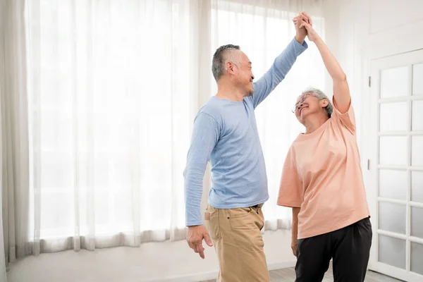 Romantic and cheerful Asian well-being senior couple enjoy dancing and holding hands to music together with smiles and happiness in the living room. Senior retirement activity at home.
