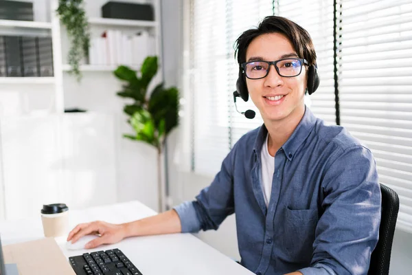 A smiling Asian man working as customer support operator with a headset in a call center. Portrait of sales agent sitting at desk and looking at camera. Customer care support service representative.