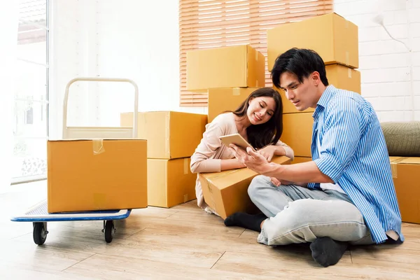 Happy Asian couple moving to a new house, Woman unboxing cardboard boxes from the old house while man online shopping things with a digital tablet. Family-Moving house relocation, home moving concept.