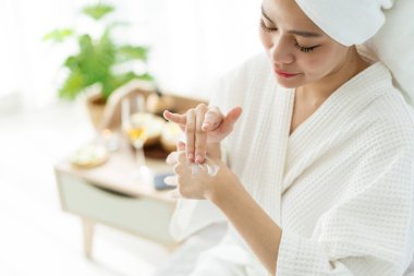 Bornozlu, nemlendirici krem sürerken ya da elinin arkasına losyon sürerken rahatlamış genç bir Asyalı kadının görüntüsü. Banyodan sonra el kremi süren bir kadın. Mevsimsel cilt koruma kavramı.