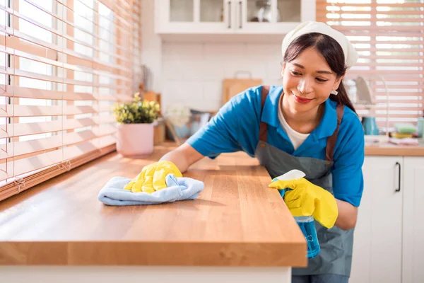 专业的亚洲妇女清洁服务 戴着黄色橡胶手套 用抹布用喷雾液清洁剂擦拭家中厨房顶部的木制柜台 打扫房间 打扫房间 免版税图库照片