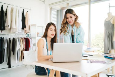 İki Asyalı moda tasarımcısı veya terzisi yeni koleksiyonun tasarımını dizüstü bilgisayarında kumaş ve çizim örnekleriyle tartıştılar. Stüdyoda birlikte çalışıyoruz..