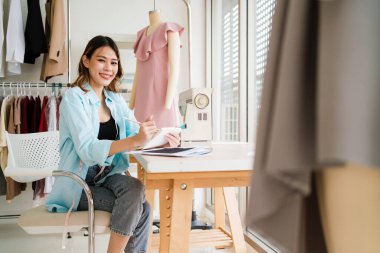 Kendine güvenen Asyalı bir terzinin portresi terzi atölyesinde giysiler üzerinde çalışıyor. Çekici bir bayan moda tasarımcısı gülümseyip arka planda elbise mankeni olan kameraya bakar..