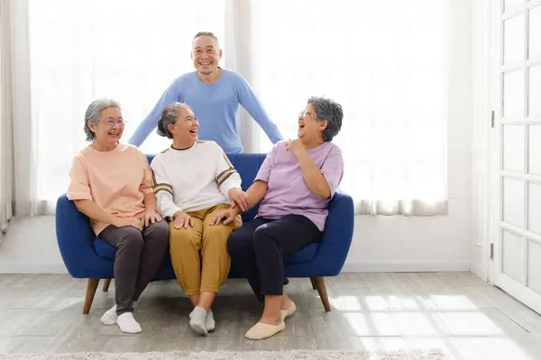 The group of happy and healthy three Asian senior women sits together on a sofa with an Asian senior man standing in the back. Smiling and laughing together. Image with copy space