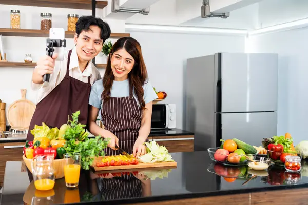 Happy Asian couple wearing aprons. Man live stream by smartphone during woman prepare a vegetarian healthy salad with fresh vegetables such as carrot, tomato cabbage and green oak in the home kitchen.