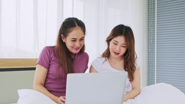 幸せなアジアの女性のカップルが一緒に笑顔と笑いで午前中にオンラインショッピングのためのラップトップを使用してベッドに横たわっている 女性はラップトップからのソーシャルネットワーク チャットやオンラインショッピングのためのデバイスを使用します — ストック動画