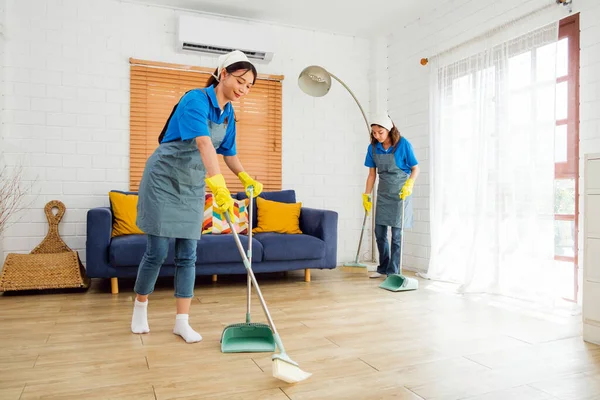 Two Asian young professional cleaning service women worker team working in the house. Girls housekeeper sweeps broomsticks on the wooden floor with another one cleaning under the sofa. Cleaner.