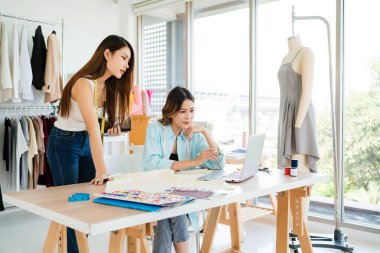 İki Asyalı moda tasarımcısı veya terzisi yeni koleksiyonun tasarımını dizüstü bilgisayarında kumaş ve çizim örnekleriyle tartıştılar. Stüdyoda birlikte çalışıyoruz..