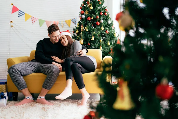 Noel Baba şapkası takan güzel, mutlu bir Asyalı kadın erkek arkadaşıyla birlikte arka planda bir Noel ağacı olan kanepede oturuyor. Kopyalama alanı olan resim.