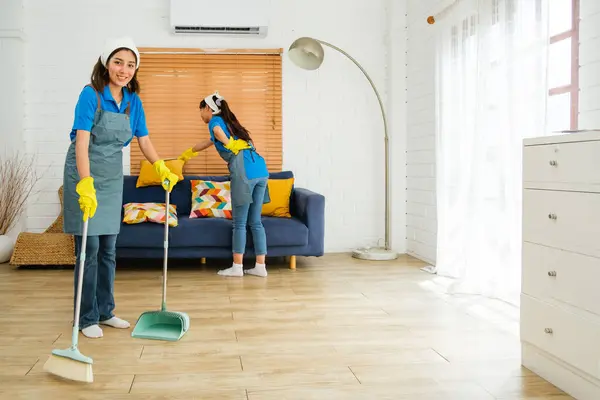 Two Asian young professional cleaning service women worker team working in the house. Girls housekeeper sweeps broomsticks on the wooden floor with another one cleaning the sofa pillow. Cleaner.
